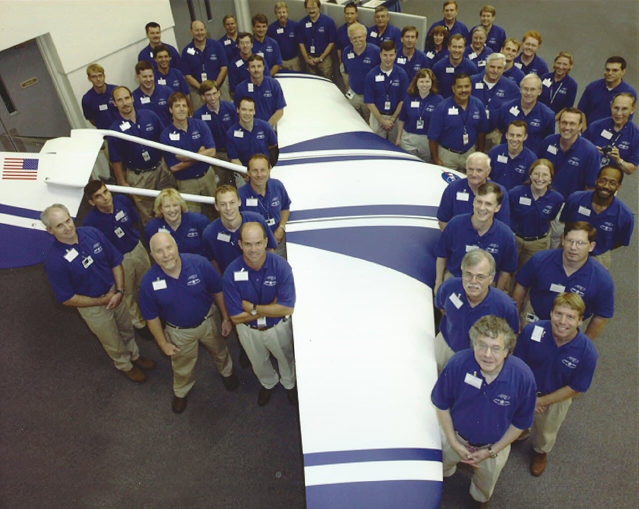 Roughly fifty members of the ARES team in matching blue ARES polo shirts, packed shoulder to shoulder around a full-scale ARES test airplane indoors at NASA Langley, the aircraft finished in white with dark blue sweeps and a United States flag on its tail fin. The Commons file page identifies team leader Joel S. Levine at the bottom right. This airframe is a ground test article built at Langley, and no ARES was ever built to go to Mars.