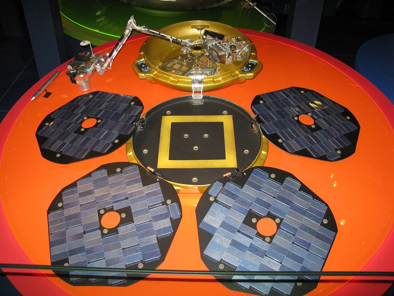 A full-size replica of Beagle 2 on display at the Science Museum in London, opened out as the lander was meant to open on Mars: the gold bowl and its instrument deck at the back, the four solar panel petals laid flat around the central lid, and the robotic arm carrying the cameras and spectrometers.