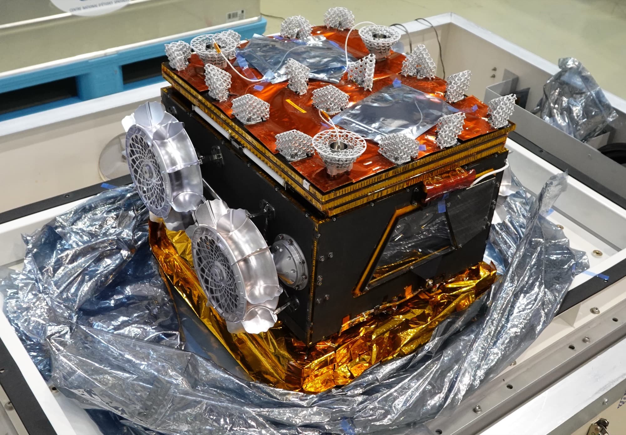 The Idefix flight rover in its cruise configuration, published by ISAS with the caption 'IDEFIX Rover' alongside the 14 March 2024 announcement of the handover to JAXA at the Mitsubishi Electric Kamakura Works. The body is wrapped in gold and black thermal blanketing, two of its four wheels are folded flat against the flank facing the camera on their stowed hinged legs, and the top face carries the orange-red blanket over the folded solar panels studded with the open aluminium lattice crash pads that take the impact when the rover is dropped. It sits in a handling frame on protective sheeting, with a Centre National d'Etudes Spatiales label visible on the wall behind.