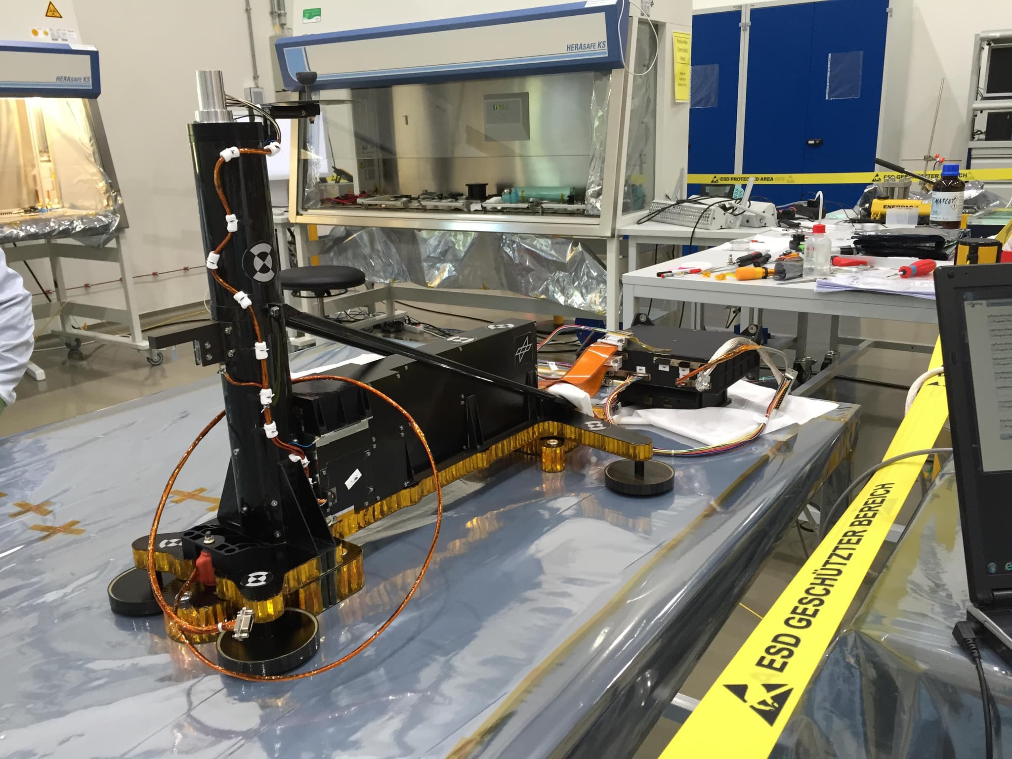 The Heat Flow and Physical Properties Package standing on a bench in a cleanroom in Germany before launch, released by JPL as PIA23044 with the caption that the heat probe was being tested in Germany. JPL does not state whether the unit shown is the flight model. The black upright tube is the support structure that holds the mole; the horizontal box beside it carries the tether compartments, and a separate electronics box sits on a cloth at the right. Yellow ESD tape reading ESD GESCHÜTZTER BEREICH runs across the foreground.