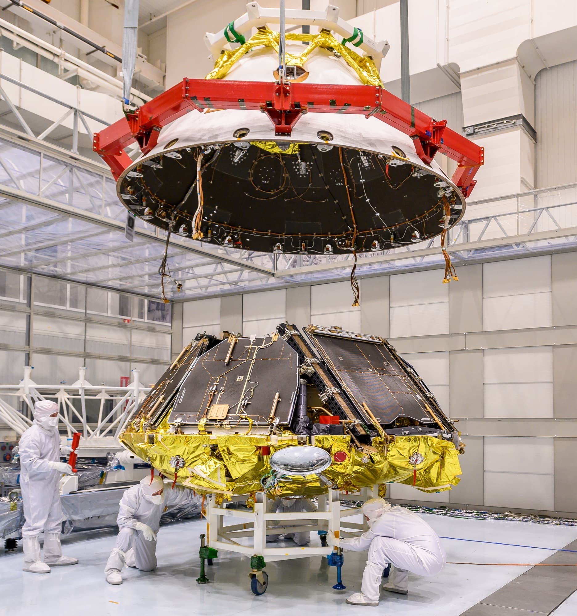 The Kazachok landing platform in the clean room at Thales Alenia Space in Turin in November 2020, sitting on a supporting trolley with its ramps and solar panels folded, while the dark rear jacket of the descent module hangs from a red lifting frame directly above it and engineers work at floor level. ESA's caption records that the front shield would later be fitted to the same trolley, and that the platform and the Rosalind Franklin rover would then undergo joint testing at Cannes.