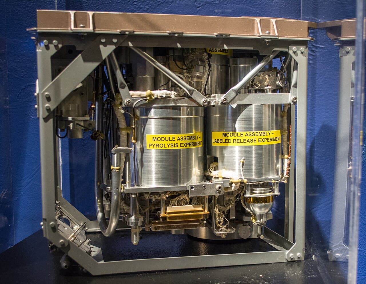 A Viking Biology Instrument in a display case at the Cleveland Museum of Natural History, photographed against a blue wall: an open grey frame holding two polished cylindrical module assemblies side by side, wired and plumbed, with yellow labels reading MODULE ASSEMBLY - PYROLYSIS EXPERIMENT and MODULE ASSEMBLY - LABELED RELEASE EXPERIMENT. The photographer's description records that the instrument was developed by TRW in Cleveland and states that this is the only one left on Earth, the other two being on Mars.