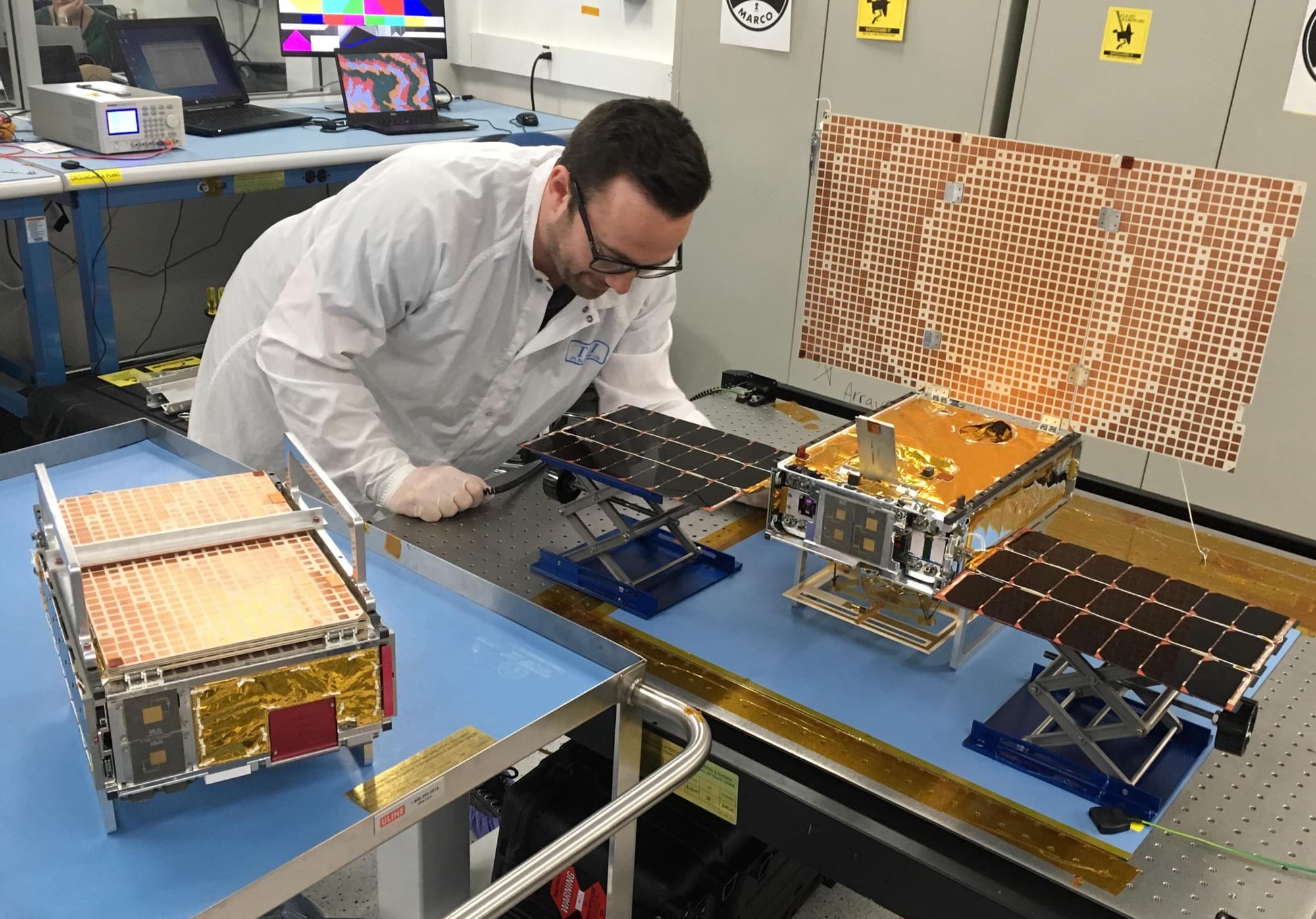 Engineer Joel Steinkraus with both MarCO spacecraft at the Jet Propulsion Laboratory in March 2018. The unit on the left is folded as it flew stowed on the rocket; the one on the right has its solar panels out, with its flat reflectarray high-gain antenna standing up behind it.