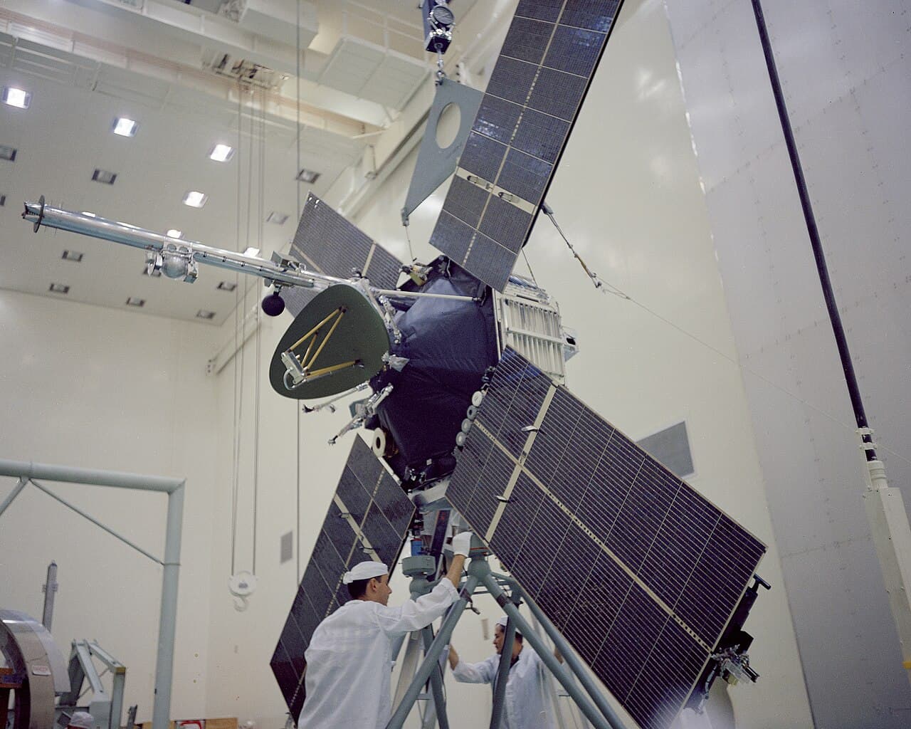 The Mariner 3 spacecraft with its four solar panels extended, being processed in the AO Building at Cape Kennedy Air Force Station on 26 September 1964 before mating with its payload fairing. Two technicians in white coats work at its base.