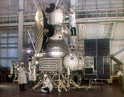 A colour photograph of an M-69 spacecraft standing in a high bay assembly hall with technicians in white coats around it and on a work platform beside it, showing the large spherical propellant tank, the plumbing loop across the body, a solar panel and the parabolic high gain antenna. Wikimedia records the subject as the verification of systems unit for Mars 1969A.