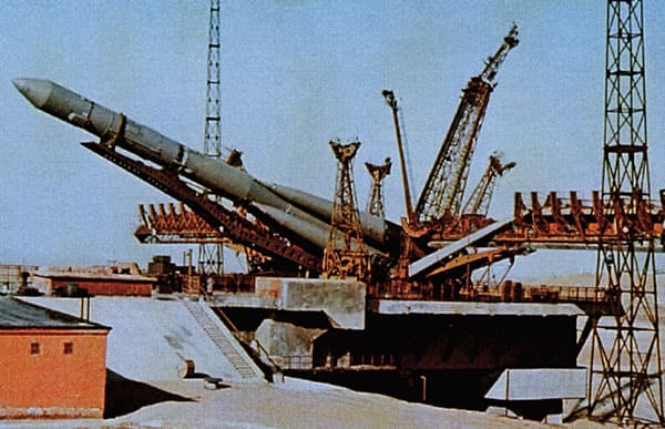 A colour photograph of a four stage Molniya 8K78 launch vehicle being raised to the vertical by its erector at Baikonur, with the service gantries, cranes and flame trench around it. The source captions the frame 'Preparing to Launch the 1M Mars Probe, Oct 10, 1960'.