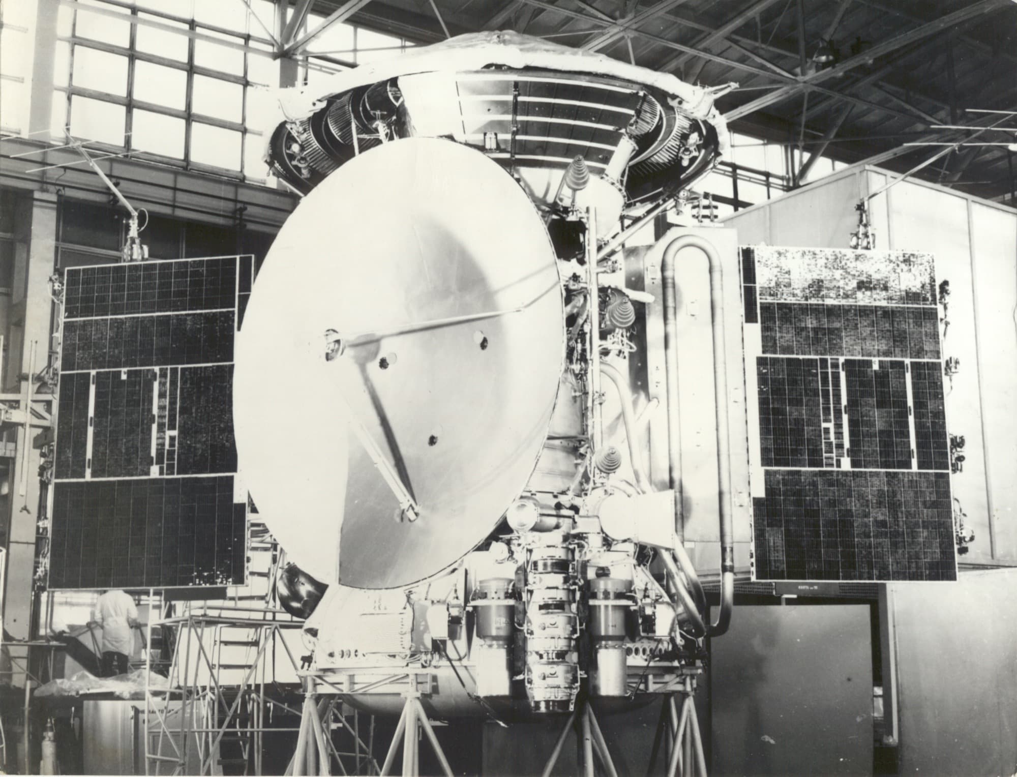 A factory photograph of an M-71 interplanetary station of the type flown as Mars 2 and Mars 3, standing on its trolley in the assembly hall with the high-gain dish and both solar wings deployed and a technician working at the left wing.