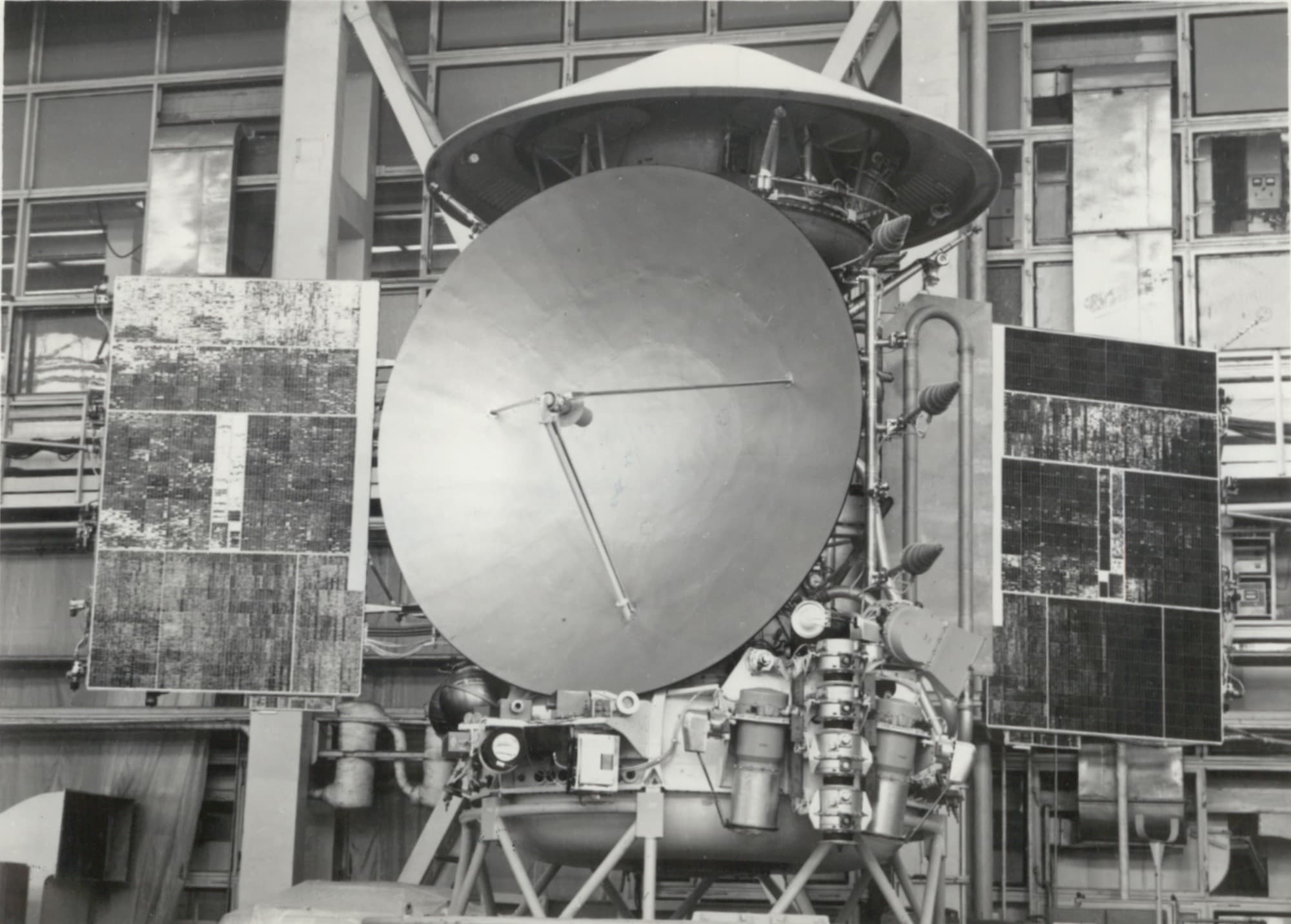 A factory photograph of an M-73 station in the assembly hall at the Lavochkin plant, solar wings and high-gain dish deployed, with the conical shroud of the upper section above the toroidal instrument compartment.