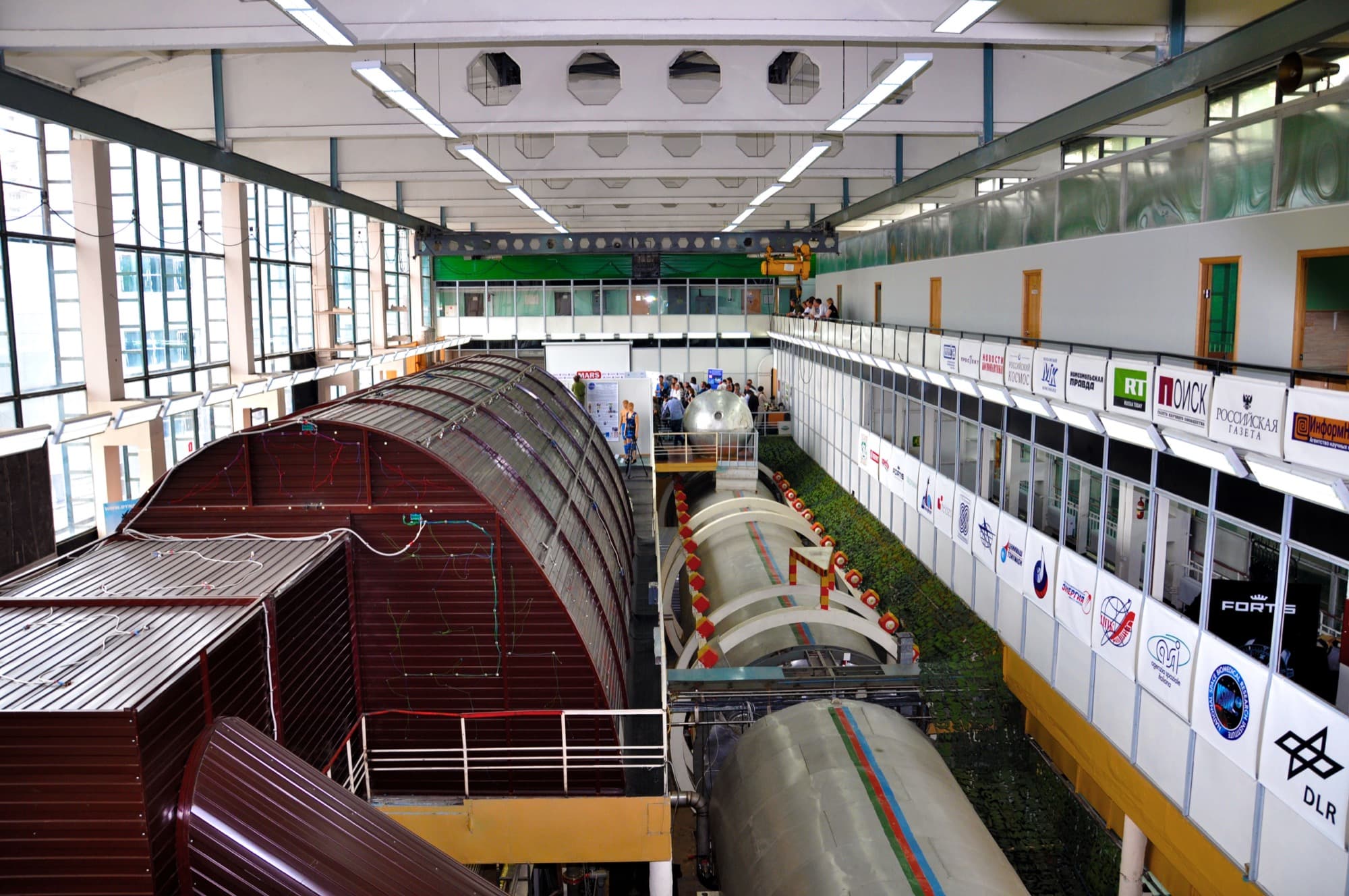 A photograph looking down the length of the NEK hall at the Institute of Biomedical Problems in Moscow, showing the linked pressure modules of the Mars500 facility from outside: a dark red barrel-vaulted module in the foreground, bare metal cylinders behind it ringed with red-painted clamps, walkways and stairs alongside, and a run of sponsor and media boards fixed to the gallery on the right.