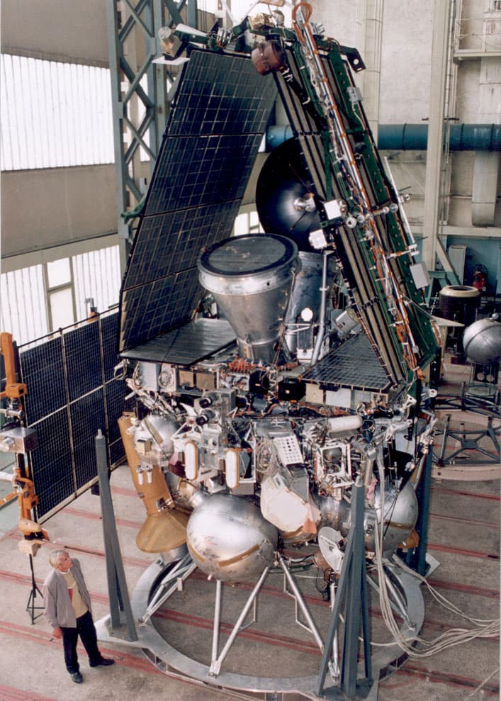 The Mars 96 orbiter standing on its handling ring in an assembly hall, solar panels partly deployed and the large spherical propellant tanks visible around its base, with a man at lower left giving the scale.