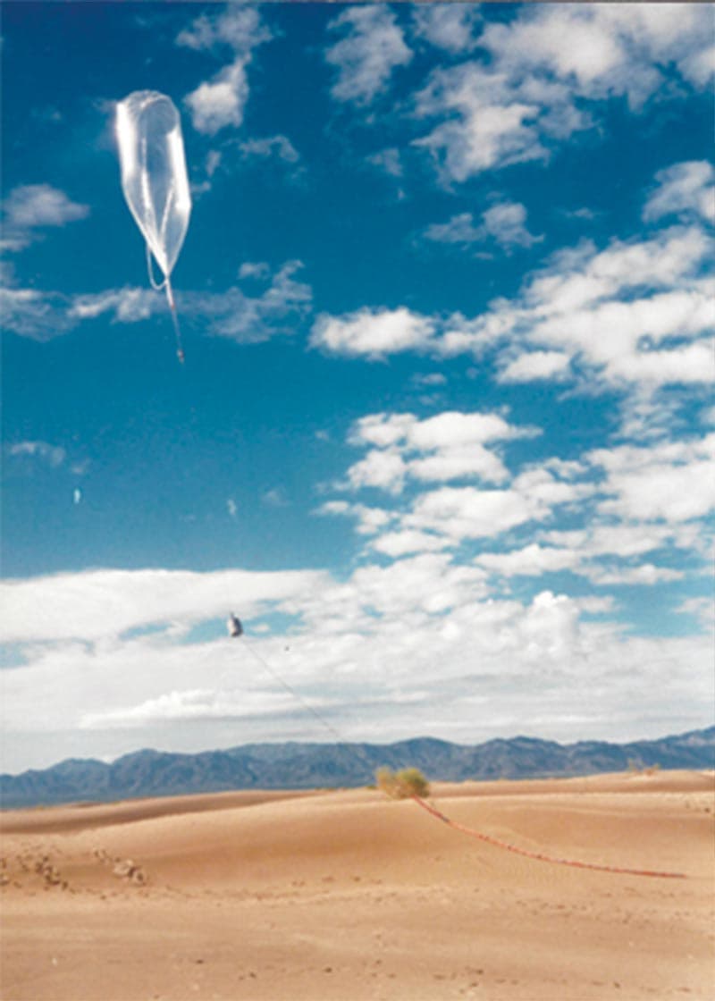 The Mars balloon in flight during the 1990 desert tests: the long envelope high against a blue sky and broken cloud, the small gondola hanging below it, and the line running down to the desert floor where the guiderope has drawn a track across a dune. The Society's timeline entry for 1990 reads: Planetary Society members support desert testing of the Mars Balloon, slated to fly on the Soviet Mars '96 mission.