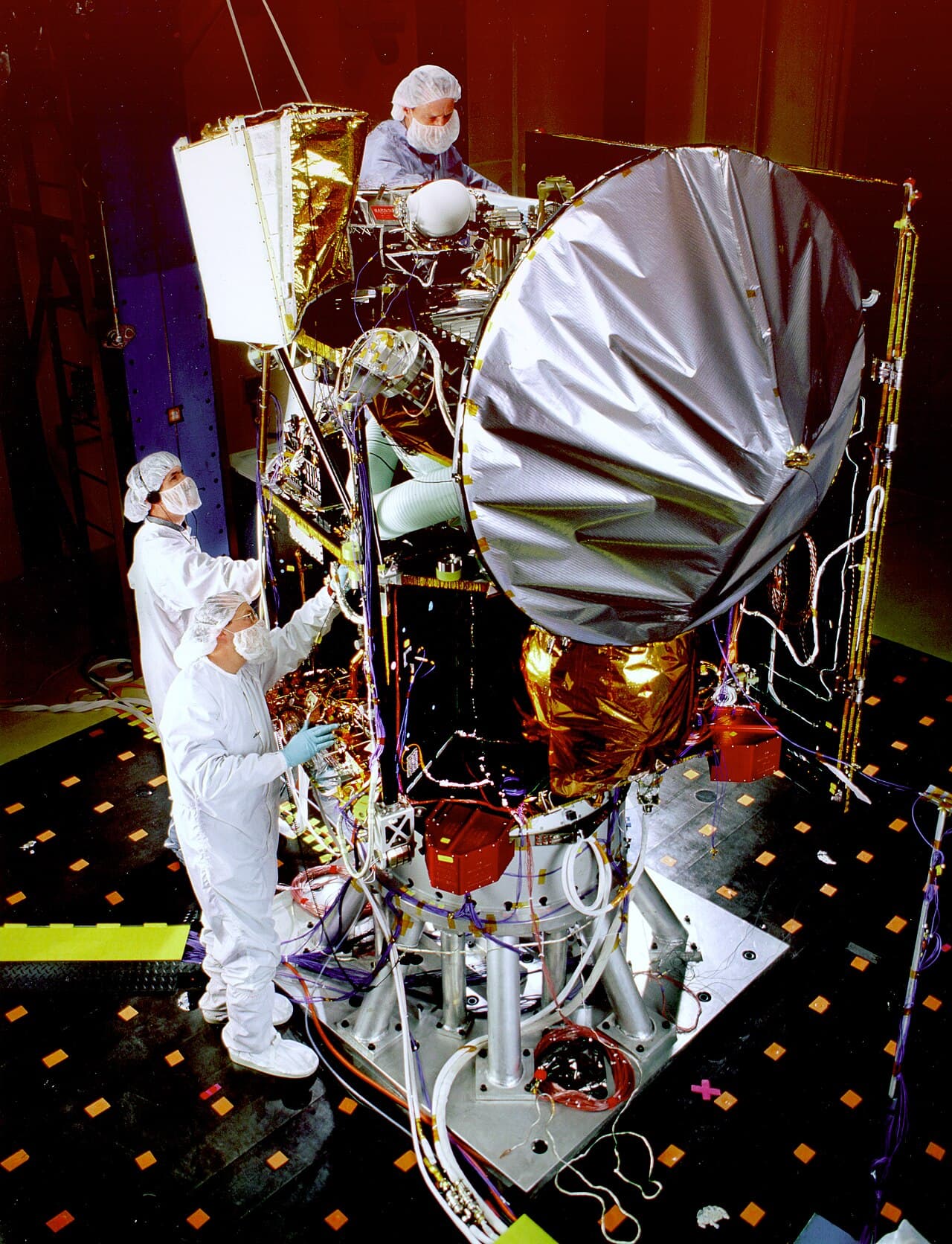 The Mars Surveyor '98 Climate Orbiter with technicians in a chamber during acoustic testing on 27 May 1998, a test that reproduces the sound levels of launch; the covered high gain antenna dish stands at the right of the spacecraft.