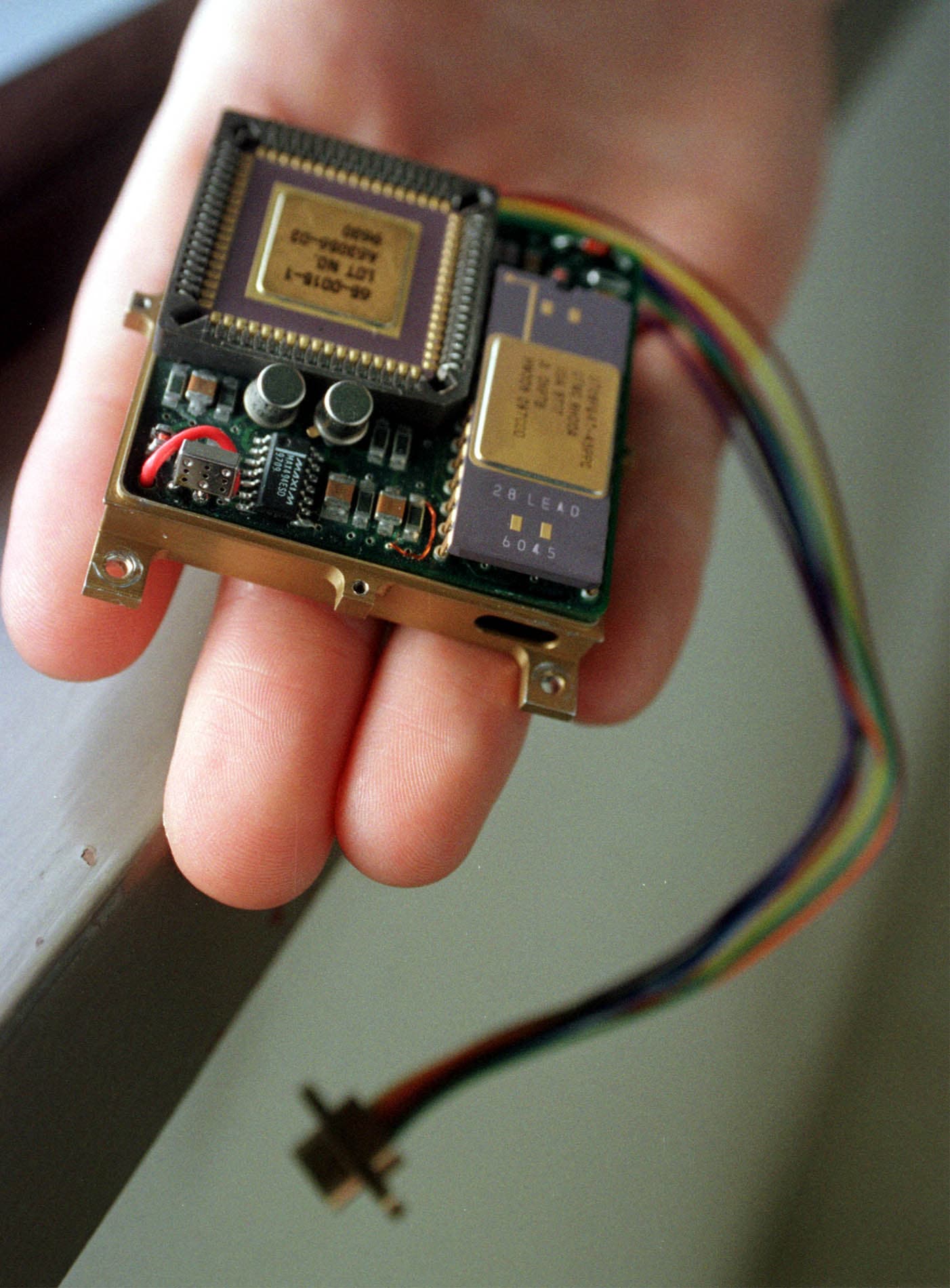 A press photograph of the Mars Microphone held flat on an open hand, showing the gold-framed board with its ceramic processor package, the two small can-shaped components, a second gold-lidded chip and a rainbow ribbon cable running off the edge. The Planetary Society captions it as the flight spare of the Society's Mars Microphone, the twin of the unit that flew aboard Mars Polar Lander.