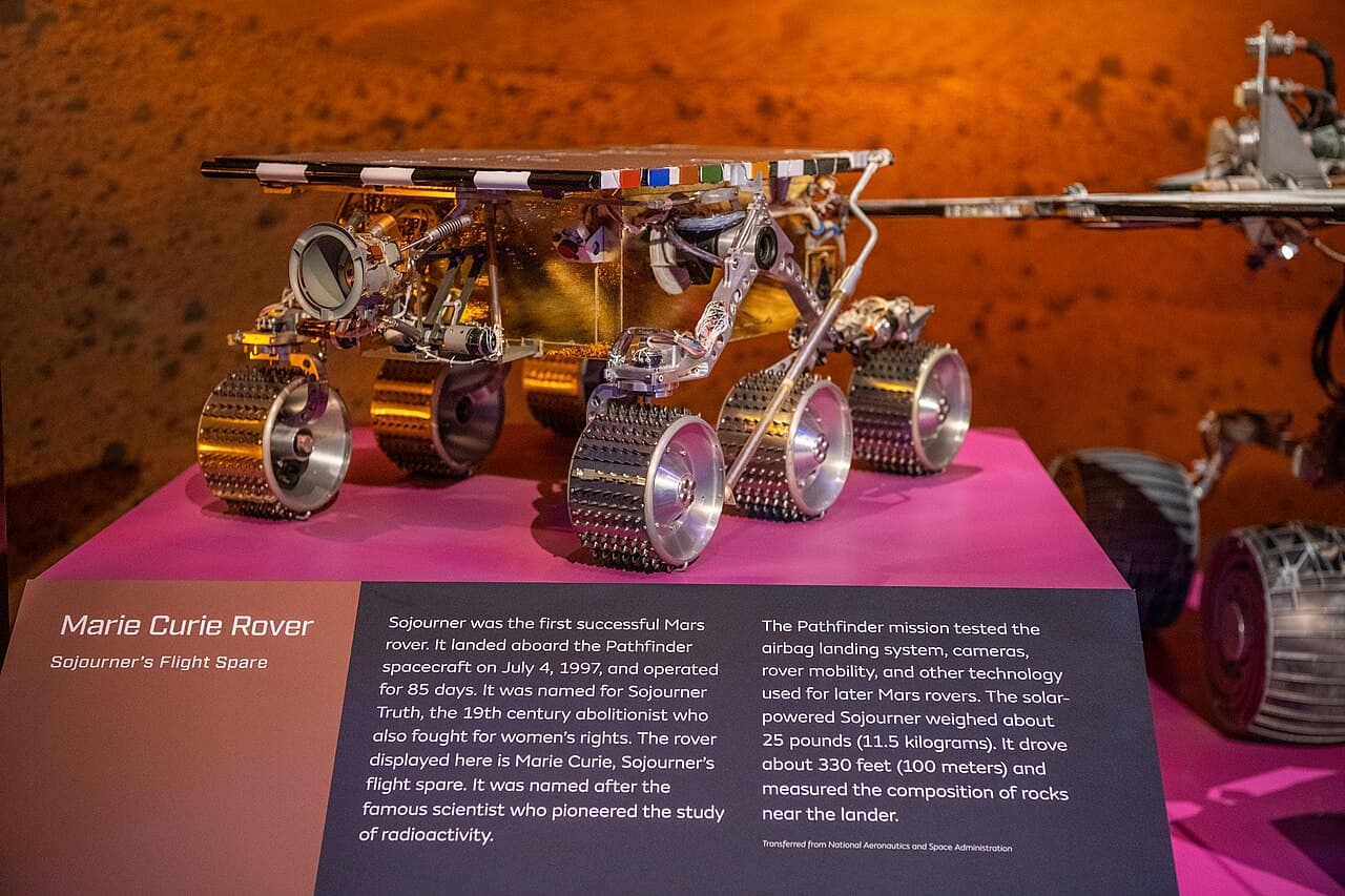 The Marie Curie rover on a magenta display plinth at the National Air and Space Museum, standing on six cleated aluminium wheels with the rocker bogie linkage and the gold warm electronics box under the flat solar panel, a strip of colour reference chips along the panel edge, and a museum label in front of it headed Marie Curie Rover, Sojourner's Flight Spare and footed Transferred from National Aeronautics and Space Administration.