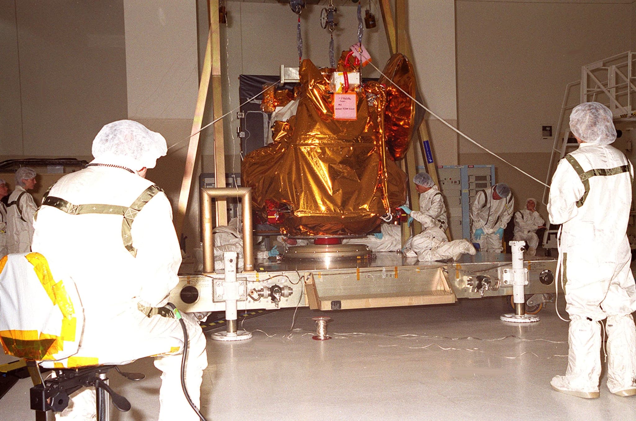 The 2001 Mars Odyssey orbiter, wrapped in gold thermal blanket and slung from an overhead crane, is lowered onto a workstand in the Spacecraft Assembly and Encapsulation Facility 2 at Kennedy Space Center on 5 January 2001 while the processing team checks its position.