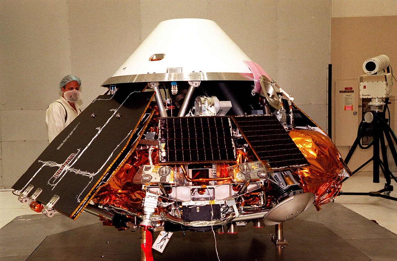 A Kennedy Space Center technician looks over the assembled Mars Polar Lander in the Spacecraft Assembly and Encapsulation Facility 2, with the white conical backshell resting above it and the solar panels spread open to either side in their deployed position.