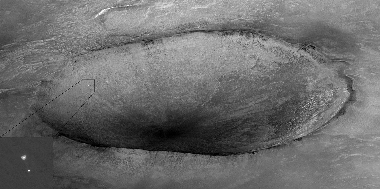 A HiRISE image from Mars Reconnaissance Orbiter showing Phoenix hanging beneath its parachute during descent on 25 May 2008, with a 10 km crater informally named Heimdall filling the frame behind it and a full-resolution inset of the parachute and lander at lower left. JPL's caption notes that although Phoenix appears to be descending into the crater it is actually about 20 km in front of it.