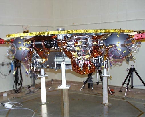A colour photograph of a Mars Surveyor 2001 lander standing on three white test stands in a bare high-bay cell, its circular equipment deck wrapped in gold and silver thermal blanketing with amber plumbing and cabling running across it, protective yellow tape and red covers on the deck edge, and camera tripods and ground support cabling on the floor around it. JPL published it in a gallery headed ATLO Test and Integration Images on the lander's own site, with no individual caption and no date.