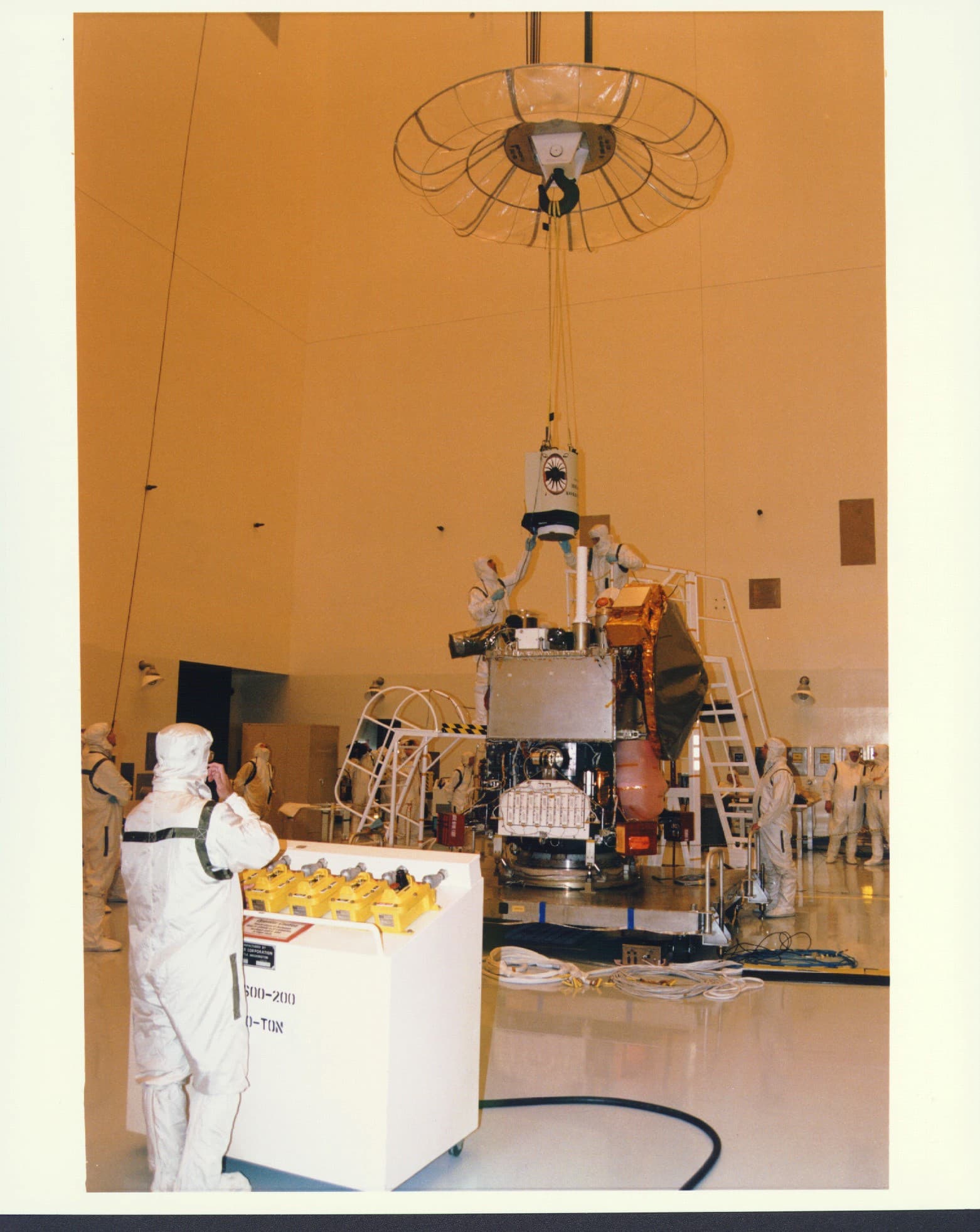 A colour photograph taken in the Payload Hazardous Servicing Facility at Kennedy Space Center on 19 August 1996, showing the Mars Orbiter Camera being lowered on a crane towards the Mars Global Surveyor spacecraft while gowned technicians on a work platform guide it into place and others watch from the floor. NASA's caption records the camera as one of a suite of six instruments and gives the launch period as opening 6 November 1996.