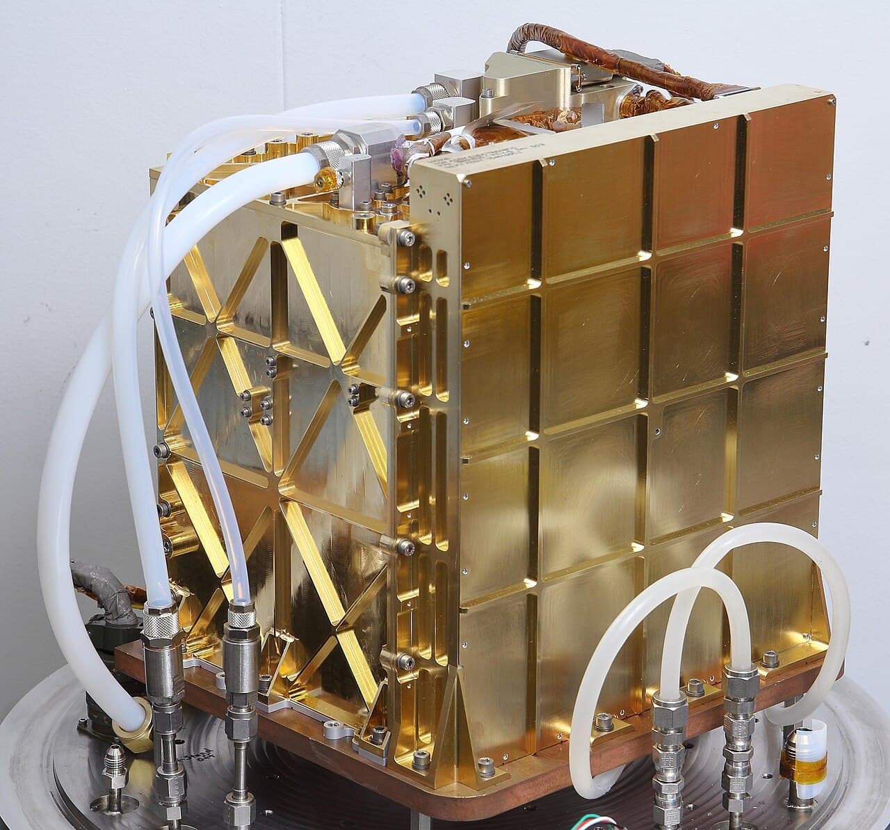 The MOXIE engineering model, the ground twin of the flight instrument, photographed on the bench at NASA's Jet Propulsion Laboratory and released on 19 January 2021. The gold-plated aluminium chassis with its machined stiffening ribs is the whole instrument: 23.9 by 23.9 by 30.9 centimetres, about 17 kilograms, with the solid oxide electrolysis unit inside it. The white PTFE tubing and the quick-disconnect fittings are laboratory test plumbing; the flight instrument carried none of it.