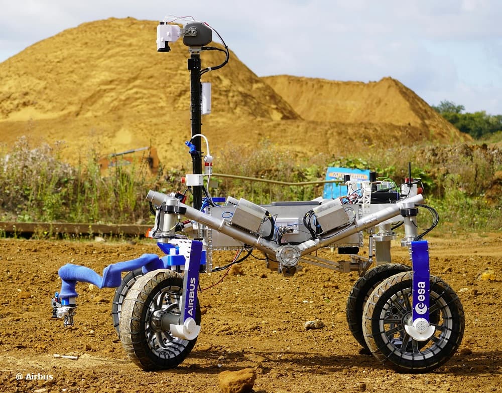 A four-wheeled Sample Fetch Rover development vehicle, nicknamed Codi and carrying Airbus and ESA markings on its wheel bogies, standing on bare excavated ground with spoil heaps behind it during ESA and Airbus field trials at Stevenage in the United Kingdom in mid-October 2023. Its blue robotic arm is extended down and forward toward a sample tube lying on the ground at the left, and a camera head and mast rise above the open tubular chassis. GMV records that the rover drove about 300 metres around obstacles and then detected and collected the tube with no human intervention, and that ESA continued the Airbus-led project after the fetch rover had been removed from Mars Sample Return.