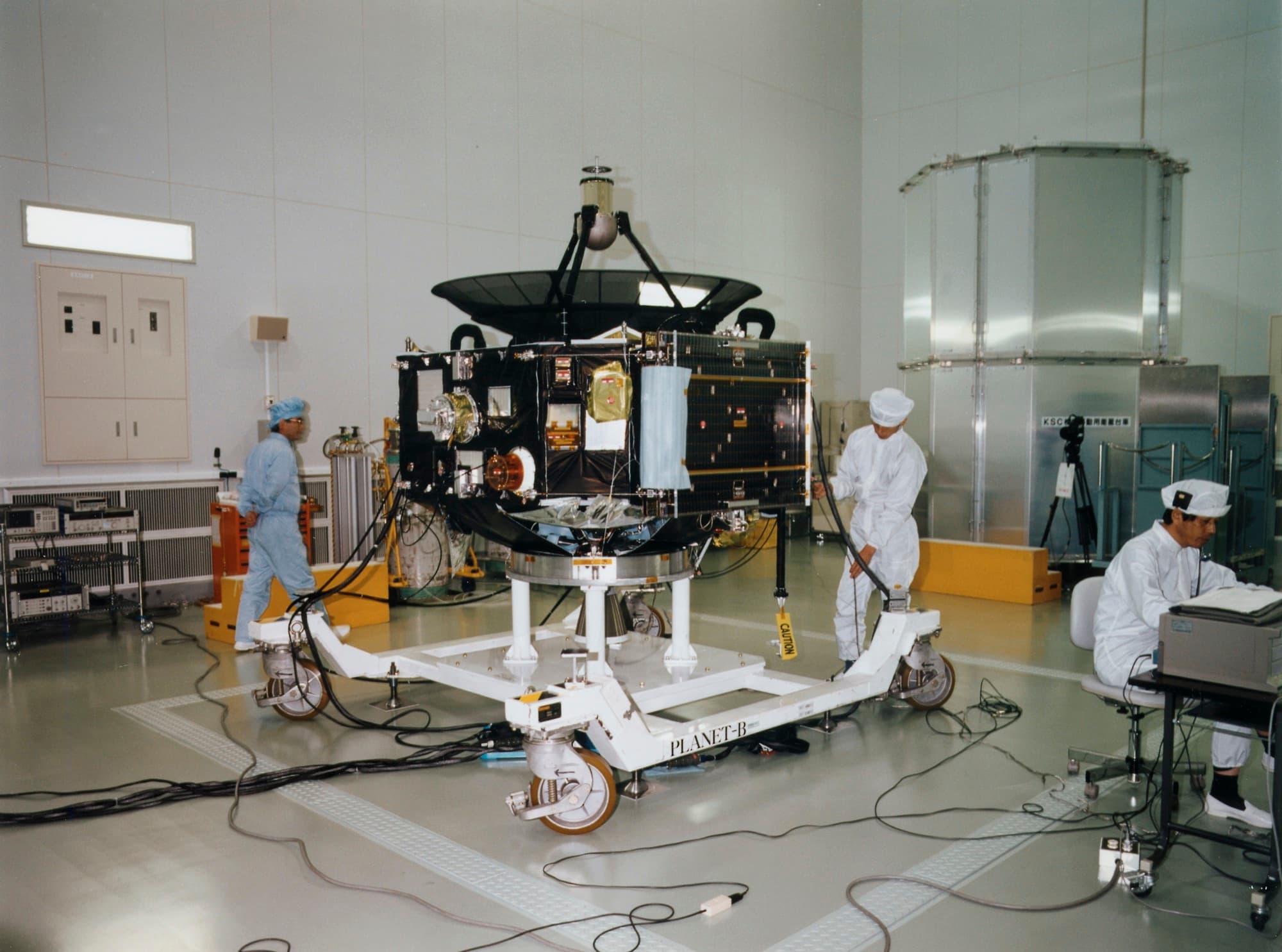 The Nozomi spacecraft standing on a wheeled handling dolly stencilled PLANET-B in an ISAS clean room, its high-gain antenna dish and stowed solar paddles visible, with three engineers in clean room suits working around it. JAXA Digital Archives item P100006474.