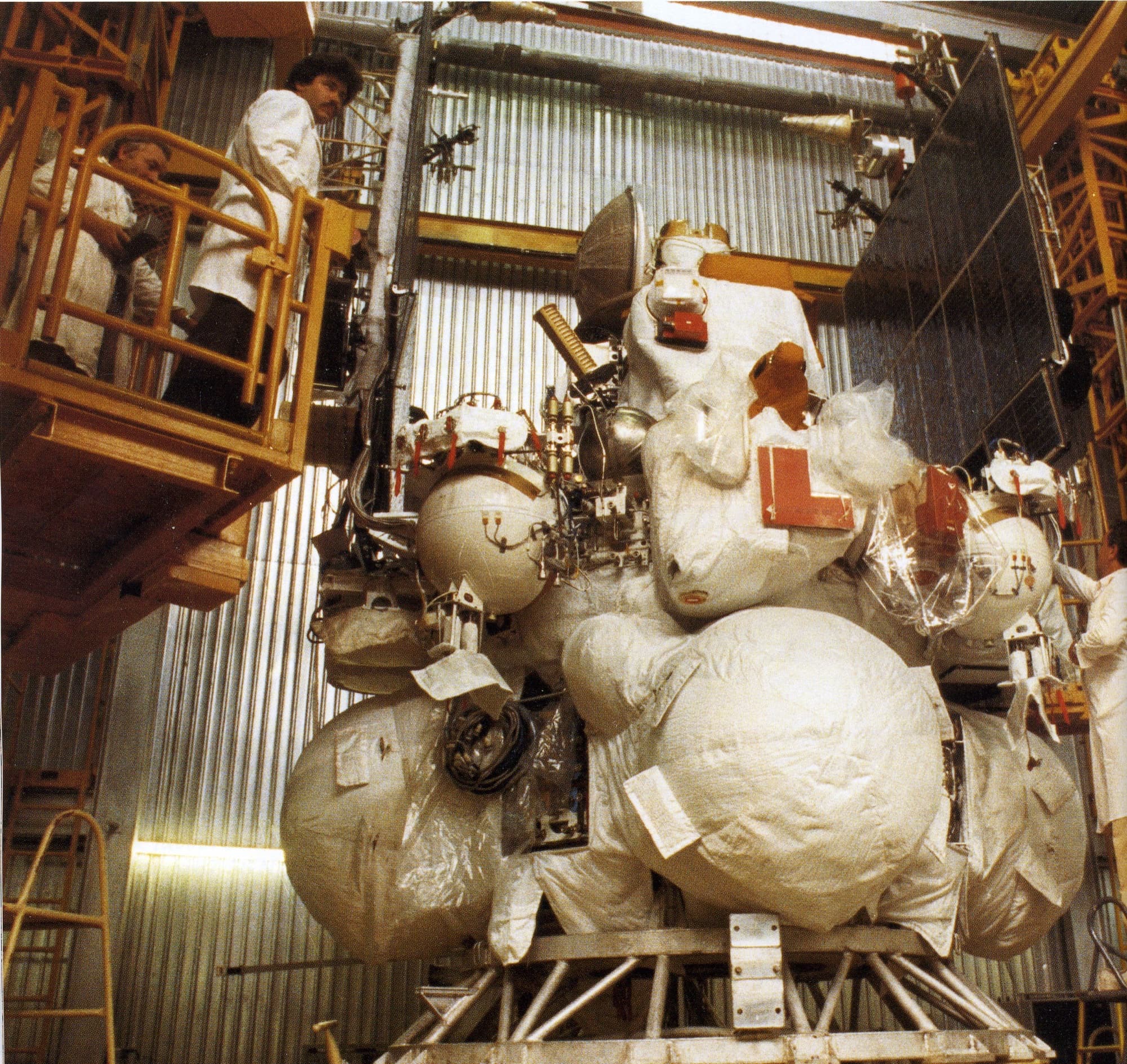 A colour factory photograph of a 1F Phobos station standing in the assembly hall at the Lavochkin plant, the thermally blanketed spherical propellant tanks of the Autonomous Propulsion Unit filling the lower half, smaller spherical tanks and instrument boxes above them, red protective covers and tags still fitted, a dark solar wing at the right edge and two technicians on a work platform at the left. The Lavochkin gallery carries no caption, date or serial number for the frame.