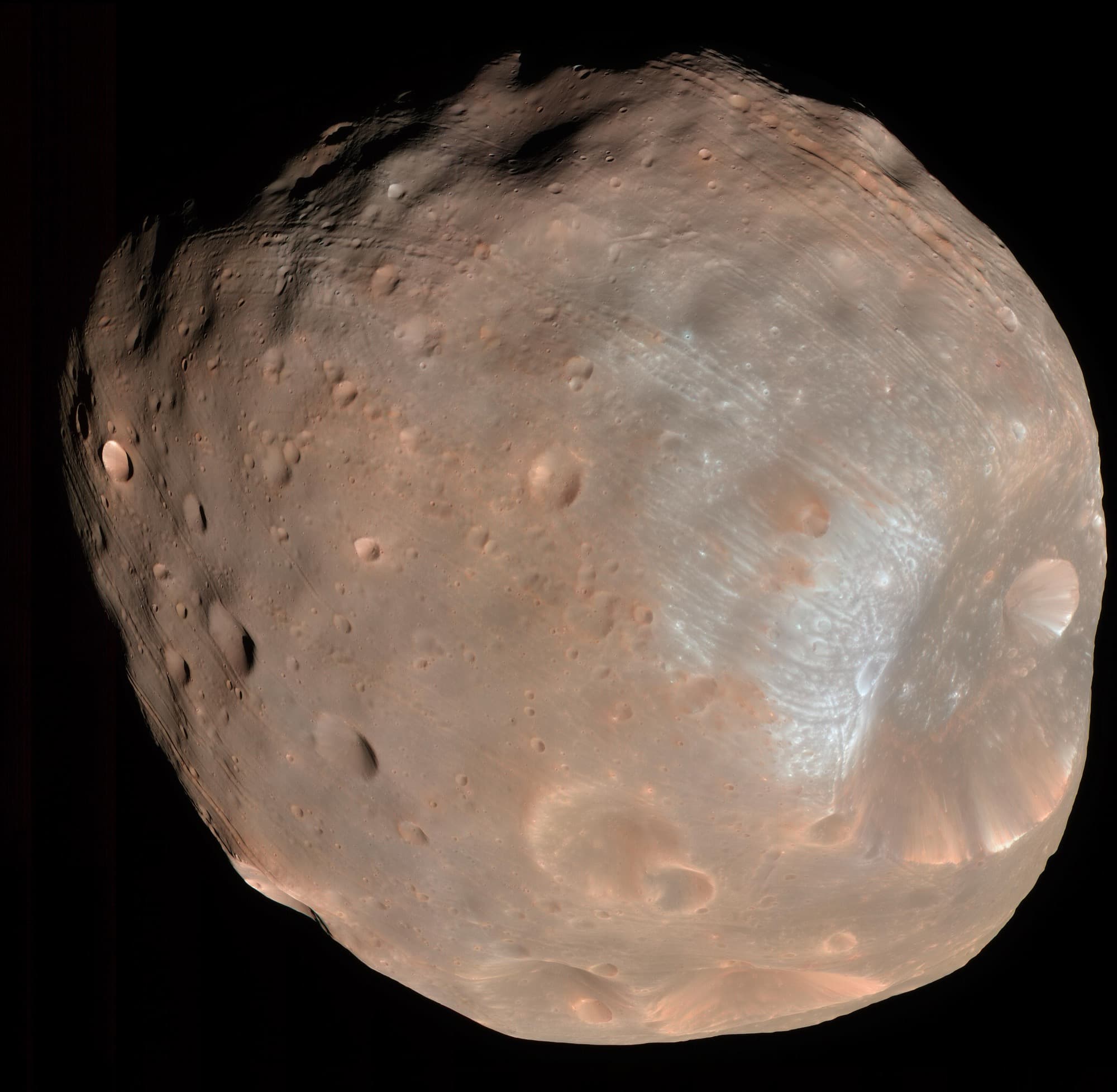 Phobos photographed by the HiRISE camera on Mars Reconnaissance Orbiter on 23 March 2008 from about 6,800 km, the first of two frames taken within ten minutes of each other. The colour is composed from the camera's blue-green, red and near-infrared channels rather than recorded as an eye would see it. JPL's caption puts Stickney, 9 km across and the largest feature on the moon, at lower right, notes that material near its rim reads bluer and so may be fresher and younger, and describes the streaked walls of the big craters as landslides of material sliding inward under a gravity less than one thousandth of Earth's. The illuminated part of Phobos in the frame is about 21 km across. JPL adds that although many of the troughs and crater chains look radial to Stickney here, Mars Express results indicate they are not related to it.