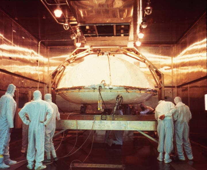 Workers in clean-room garments stand around a Viking lander capsule sealed inside its white bioshield, on its handling frame in the terminal sterilisation chamber at Kennedy Space Center in 1975. NASA's caption for this photograph reads: "Workers at NASA's Kennedy Space Center prepare the Viking 1 lander encased in its bioshield for its sterilization." NASA's 2024 Planetary Protection Handbook prints the same photograph as its Figure 2, captioned "Mars Viking landers were sterilized at KSC in ovens built specifically for the spacecraft." The first capsule went into the chamber on 15 June 1975 and spent more than 43 hours in heated nitrogen reaching 116.2 C; the second followed for almost 50 hours.