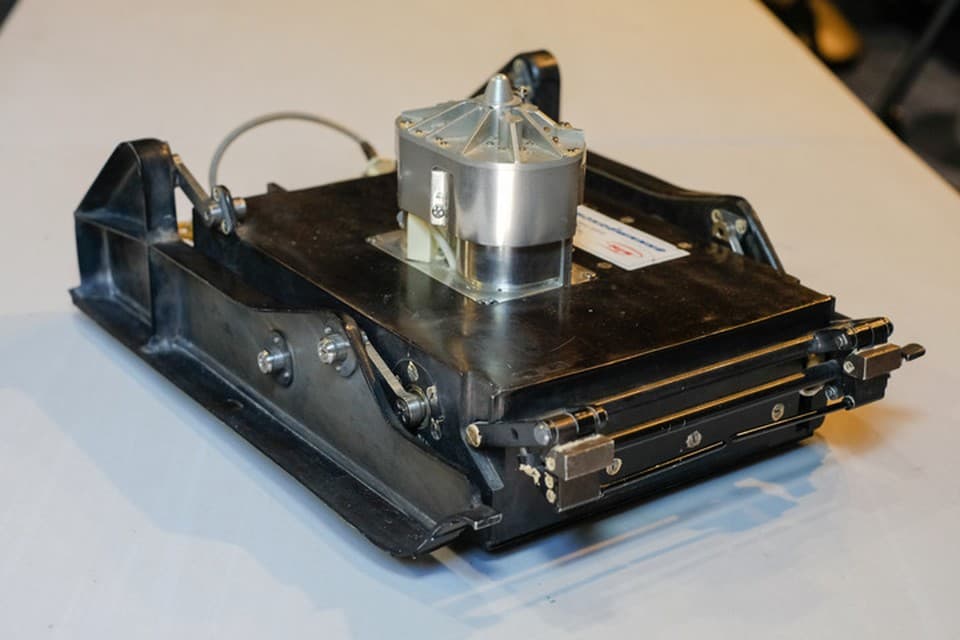 A colour studio photograph of a surviving PrOP-M laid on a white surface, showing the flat black body, the wide flat ski down each side, the aluminium penetrometer turret bolted to the top deck and the pair of thin obstacle rods projecting from the far end; Komsomolskaya Pravda's caption reads that the world's first Mars rover was only the size of a large book, and its article states that three Leningrad-built units survive, one at NPO Lavochkin, one at VNIITransMash and one on show in the Museum of Cosmonautics in the Peter and Paul Fortress, without tying this studio frame to any one of them.