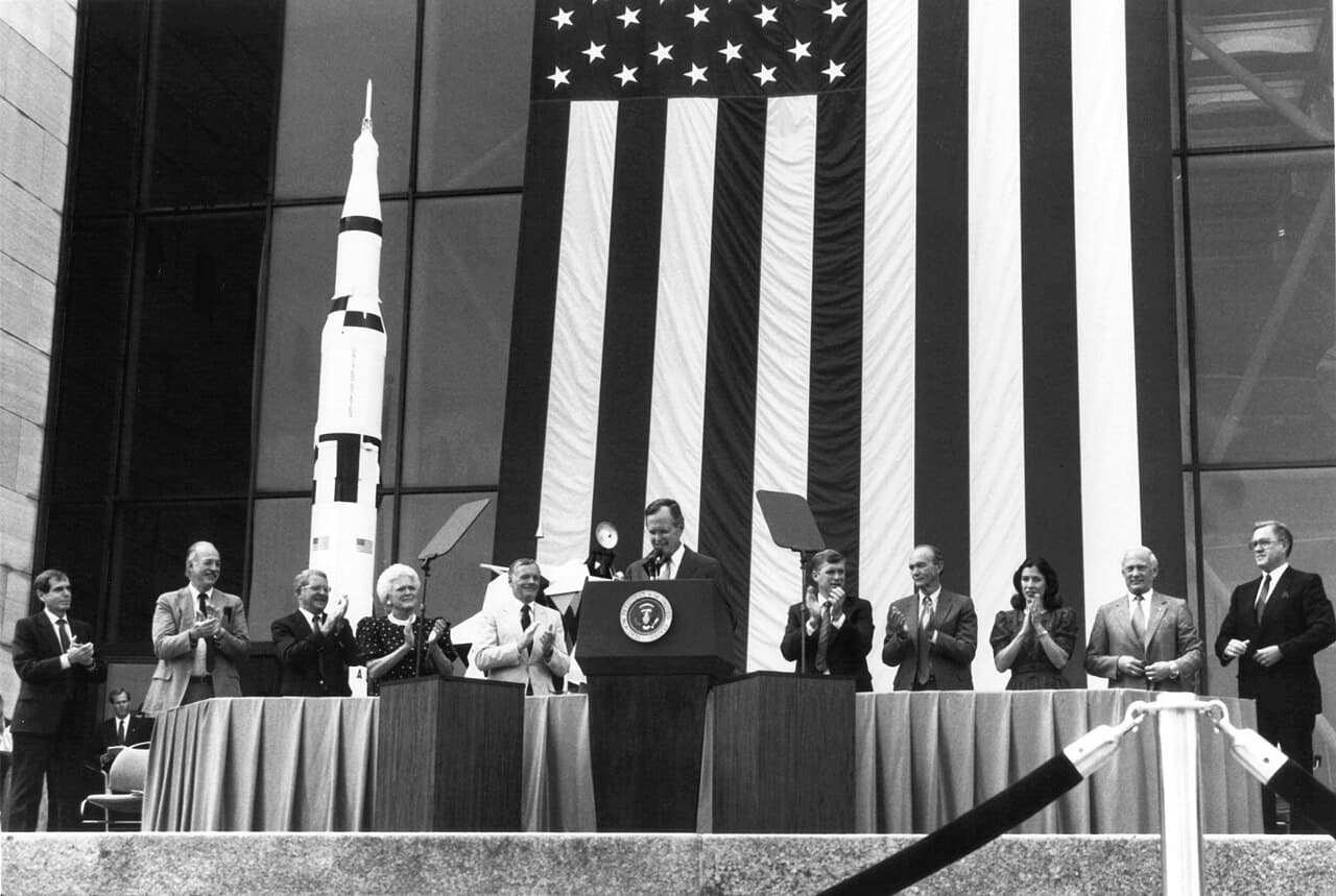 A black and white photograph of President George H. W. Bush speaking from the presidential podium on an outdoor stage at the National Air and Space Museum in Washington on 20 July 1989, at the ceremony marking the twentieth anniversary of the Apollo 11 landing. A very large United States flag hangs behind the stage with a Saturn V model standing beside it, and the platform party, which included the Apollo 11 crew, Vice President Dan Quayle and NASA Administrator Richard Truly, is applauding. NASA's own caption records that this is where Bush announced the Space Exploration Initiative.