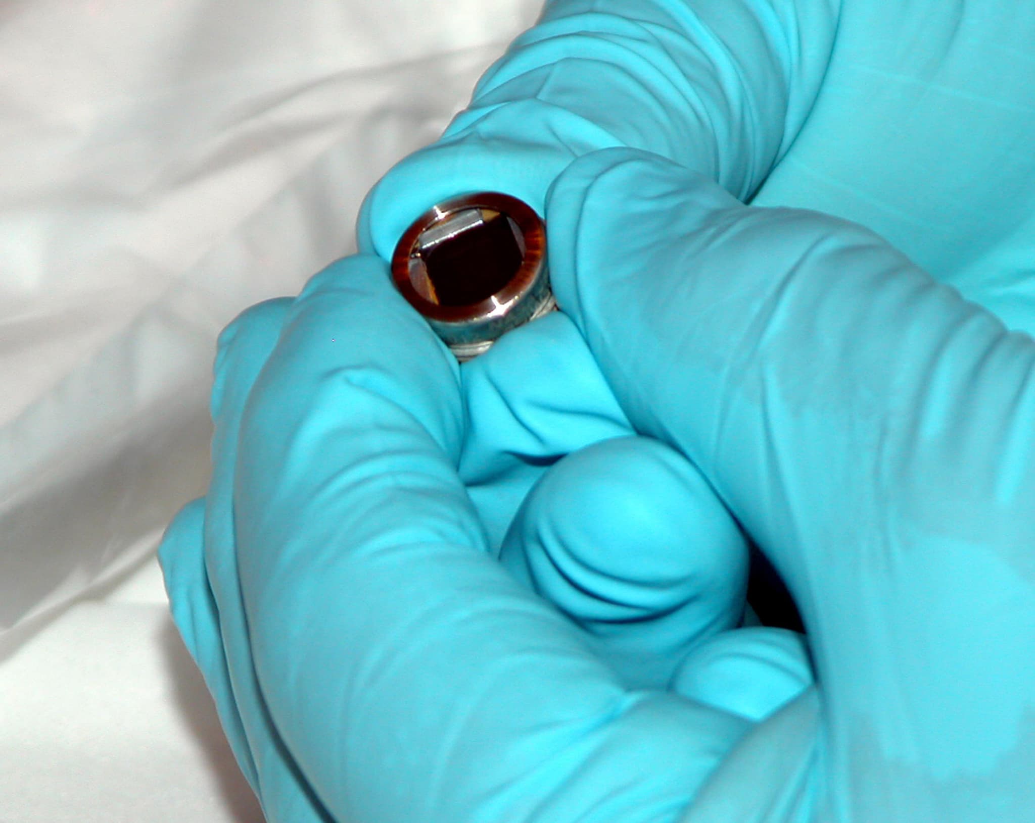 A close-up of a gloved hand holding the small mounted silicon chip that carries about 35,000 laser-engraved signatures collected from visitors to the Mars Exploration Rovers at the Jet Propulsion Laboratory, photographed before the chip was fitted to the second rover.