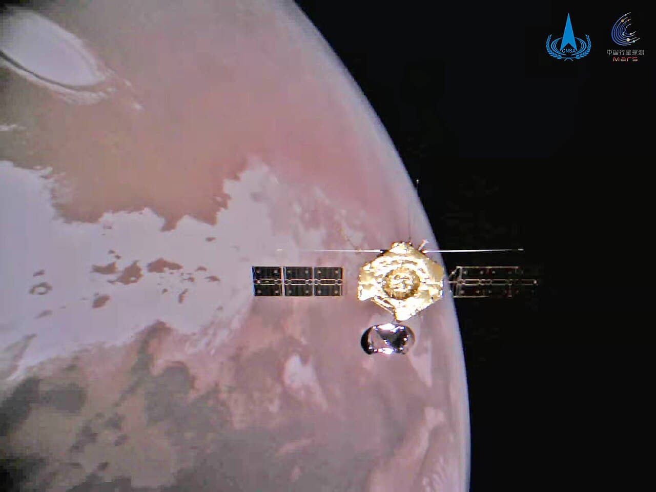 The Tianwen-1 orbiter photographed in Mars orbit by a small camera the spacecraft released, showing the gold-foiled bus, both solar wings, the high-gain antenna and the subsurface radar booms, with Mars filling the frame behind it and the bright north polar cap at upper left.