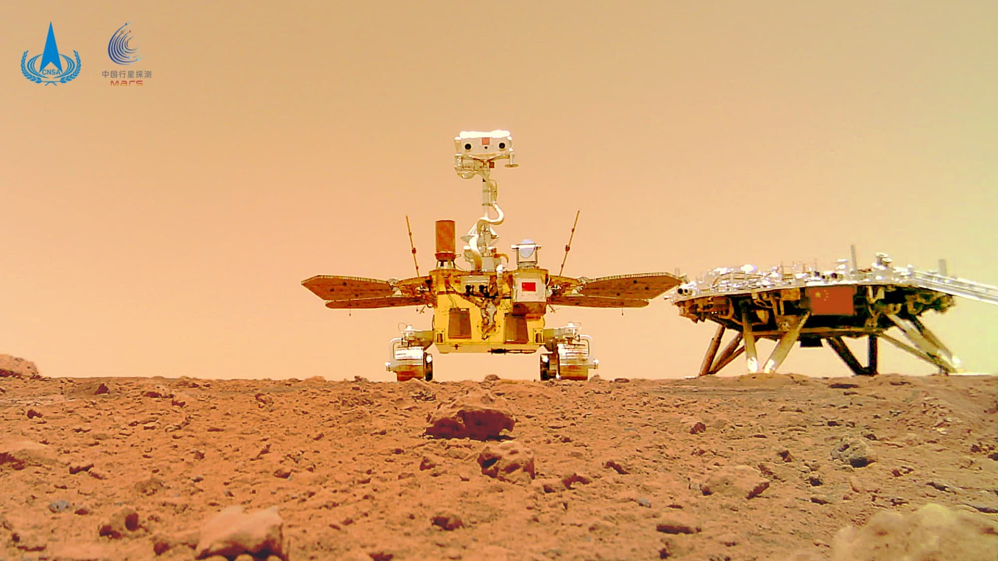 The image CNSA publishes as “着巡合影”图, the lander and rover group photo, shot from ground level with the Zhurong rover at the left, solar wings and mast deployed and a red flag on its body, and the landing platform at the right. CNSA's own text states that the rover drove to about 10 m south of the landing platform, released the separation camera mounted on its underside, reversed back towards the platform, and that the camera photographed the rover moving and then the rover and platform together, the frames going by radio to the rover and on to Earth through the orbiter relay.