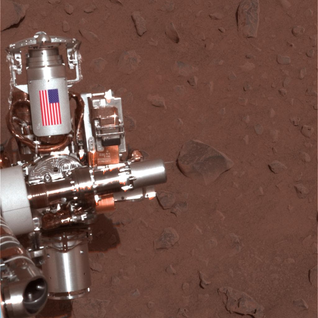 A Spirit panoramic camera image taken on 2 February 2004, sol 30, showing the rover's arm turret against the ground of Gusev crater, with the cylindrical cable guard of the rock abrasion tool at the upper left carrying an image of the United States flag. The shield was machined from aluminium recovered from the World Trade Center site.