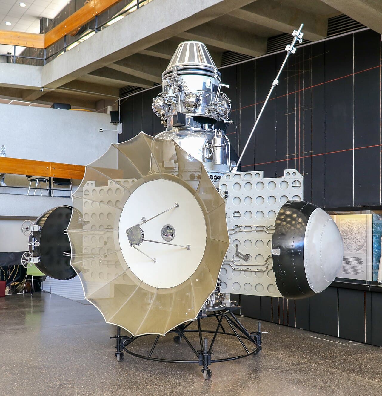 A 3MV station exhibited as Zond 3 in the hall of the Tsiolkovsky State Museum of the History of Cosmonautics in Kaluga: the petal-built parabolic dish across the front, two solar wings, the polished pressurised instrument compartment with its spherical tanks and orientation sensors above, the two hemispherical thermal radiators at the sides, and a wheeled museum stand under the whole thing.